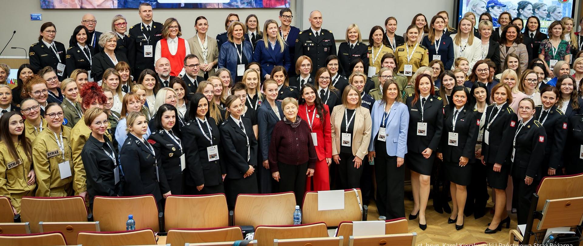 Grupa uczestniczek i uczestników konferencji pozująca do wspólnego zdjęcia w auli Akademii Pożarniczej. Wśród nich liczne funkcjonariuszki PSP, Policji i innych służb oraz przedstawicielki instytucji cywilnych. W tle ekran z napisem dotyczącym konferencji „Kobiety w systemie bezpieczeństwa państwa”.