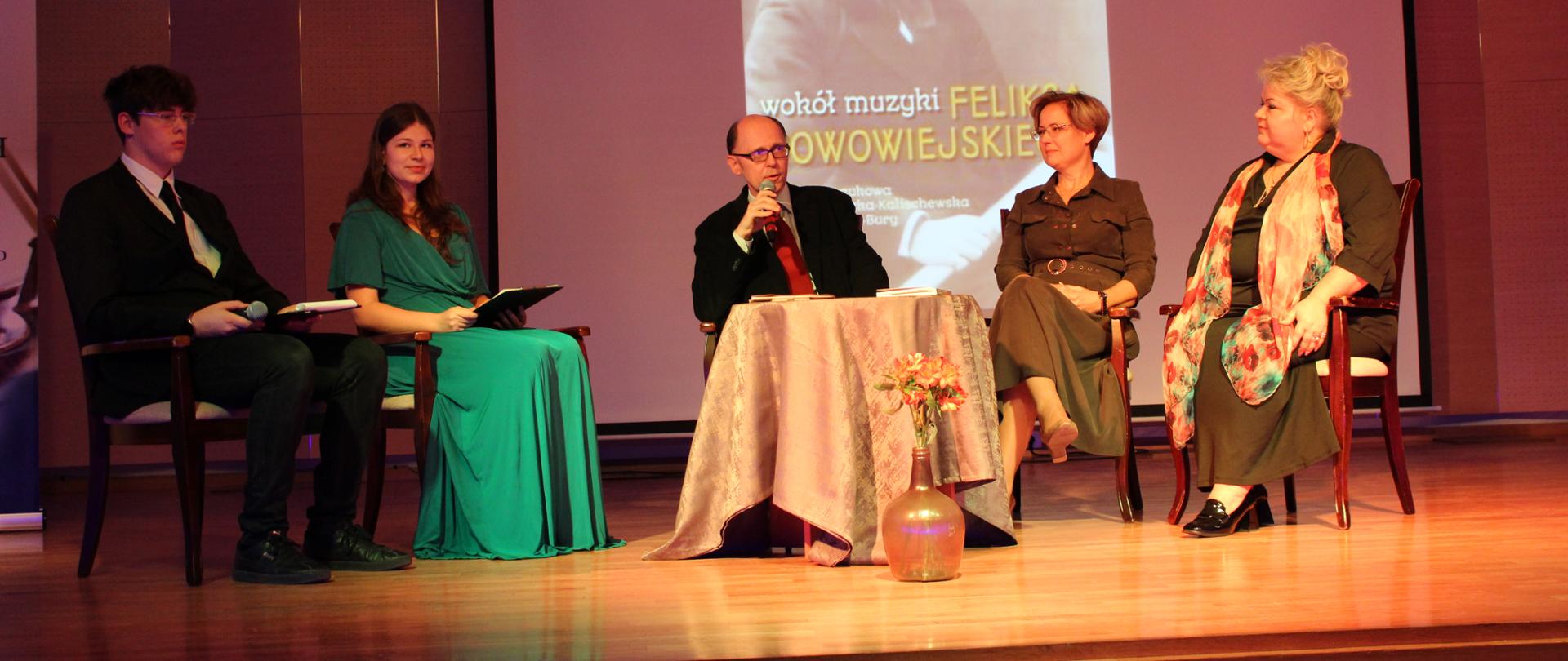 zdjęcie przedstawia organizatorów spotkania podczas wykładu na krzesłach przy stoliku na scenie sali koncertowej, w tle ekran multimedialny z portretem bohatera wykładu