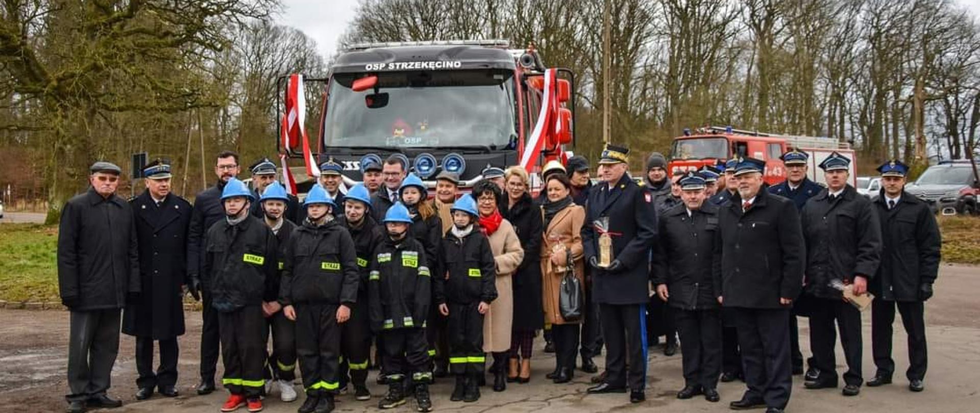 Zdjęcie grupowe z uroczystości, w tle nowy samochód ratowniczo gaśniczy.