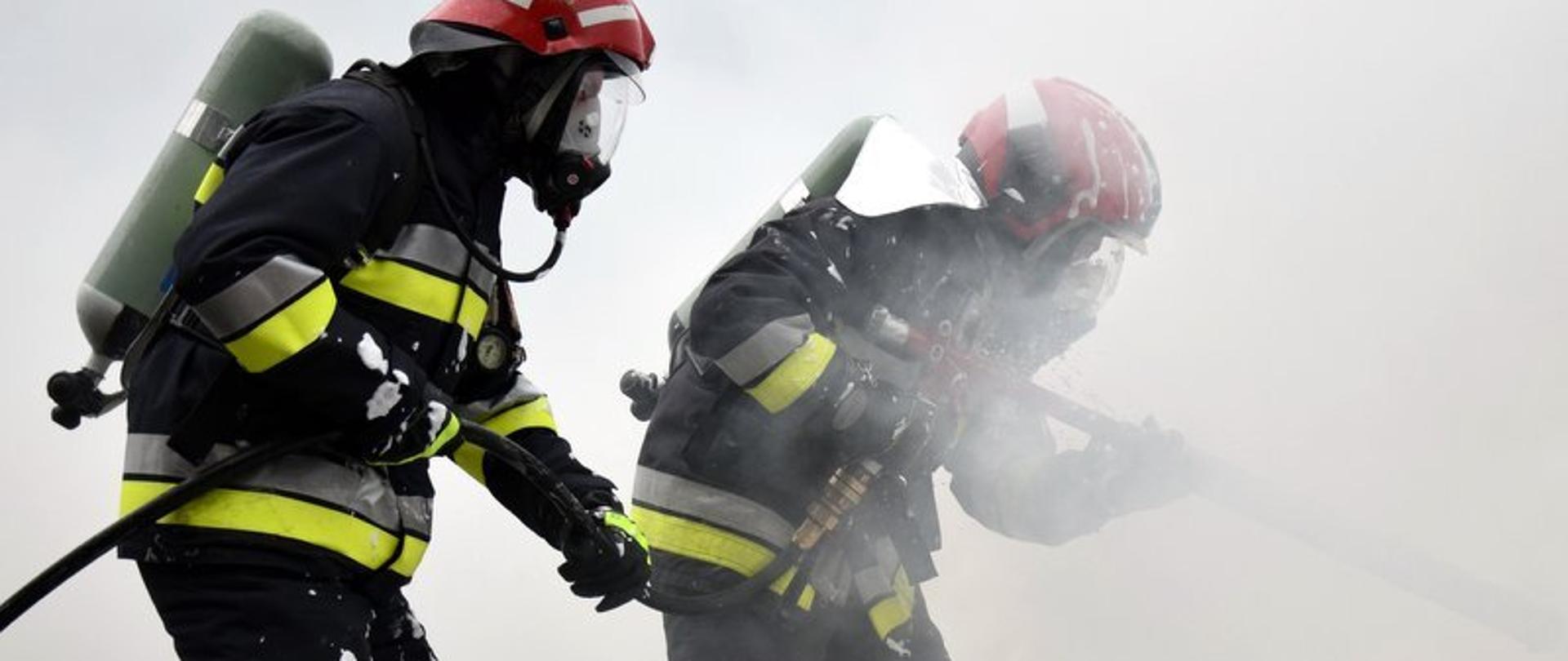Dwóch strażaków w zadymionym miejscu gasi pożar. Strażacy w ubraniach specjalnych z aparatami powietrznymi.