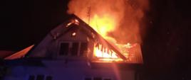Zdjęcie przedstawia palący się dach murowanego domu. Z dachu widać wydobywające się płomienie ognia i unoszący się dym. Przednia część dachu jest uszkodzona. Jest pora nocna. Prze budynkiem widać stojącego strażaka w ubraniu specjalnym i hełmie. Przed budynkiem widać cześć ogrodzenia.