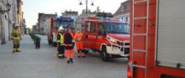 Pożar kamienicy na rawickim rynku. Na ulicy stoją trzy wozy strażackie. Auta mają włączone niebieskie światła błyskowe. Przy mniejszym samochodzie znajduje się załoga zespołu ratownictwa medycznego oraz trzech strażaków. W tle budynki.