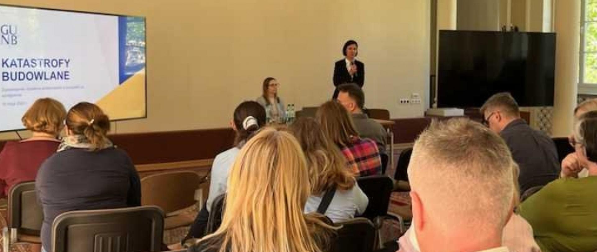 Osoby uczestniczące w prezentacji na temat katastrof budowlanych; prelegent przy ekranie z napisem „GUNB Katastrofy Budowlane”.