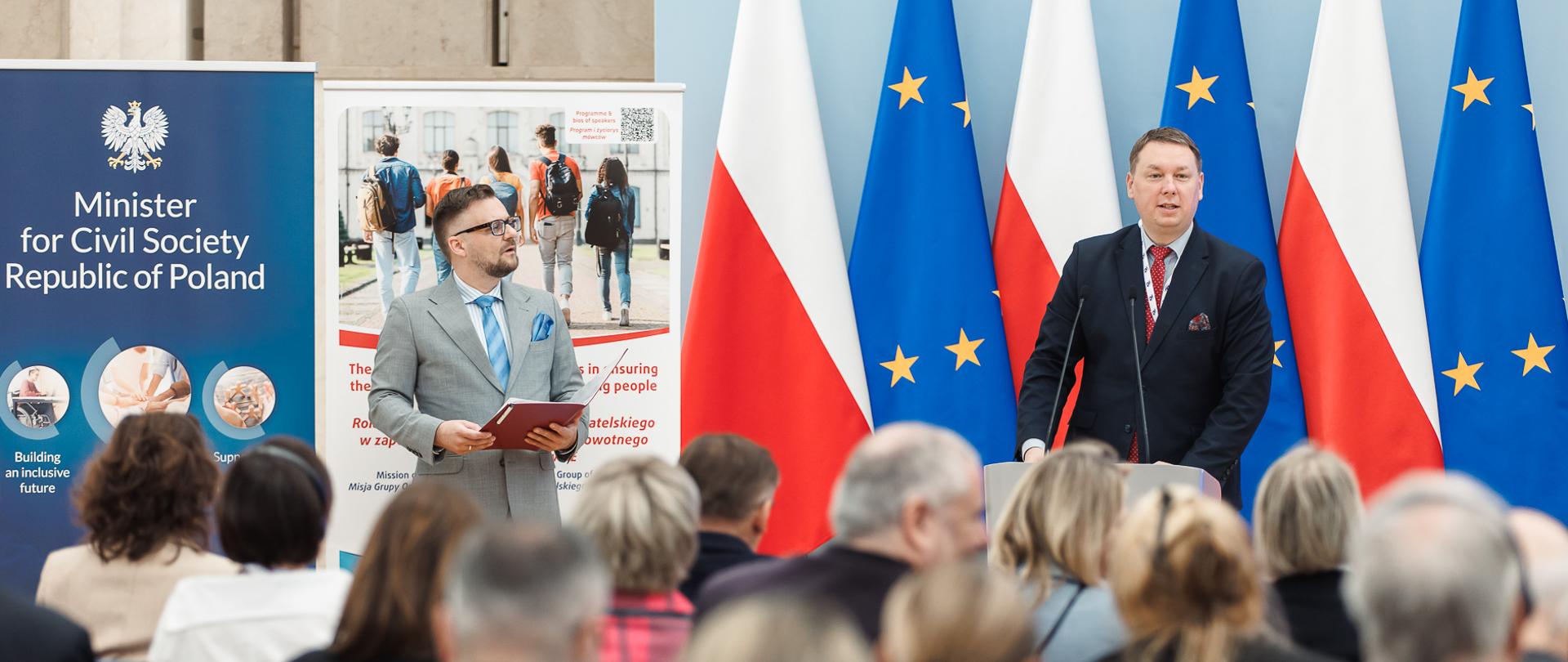 Zdjęcie pokazuje uczestników konferencji „Rola organizacji społeczeństwa obywatelskiego w zapewnieniu bezpieczeństwa zdrowotnego dzieci i młodzieży w UE”, która odbyła się 15 maja 2025 r. w KPRM. Szeroki plan. Wiceminister Mariusz Krawczyk przemawia podczas sesji otwarcia konferencji. Na pierwszym planie widać sylwetki gości zgromadzonych na widowni. W tle widoczne flagi Polski i UE oraz ekran, na którym wyświetlony jest tytuł wydarzenia.
