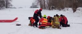 W zimowym, zaśnieżonym terenie grupa ratowników pochyla się nad osobą leżącą na śniegu. Wszyscy mają na sobie jaskrawe, dobrze widoczne ubrania ochronne: czerwone, żółte i pomarańczowe kombinezony oraz kaski. Na plecach części z nich widać napis „STRAŻ”. Obok leży czerwone sanie ratownicze, które mogą służyć do transportu poszkodowanego. W tle stoją nagie, zimowe drzewa i rozciąga się spokojny, wiejski krajobraz. Cała scena wygląda jak akcja ratunkowa prowadzona w trudnych, mroźnych warunkach.