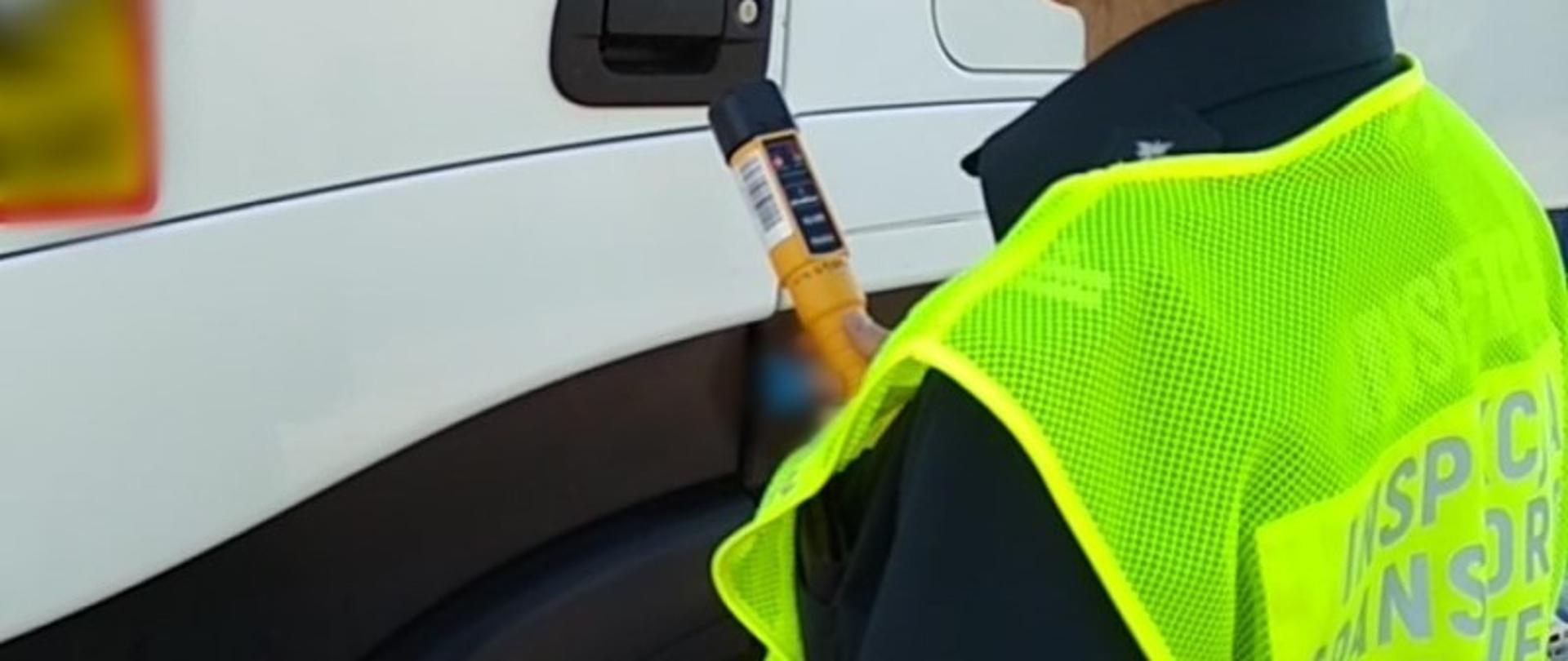 Inspektor ITD w odblaskowej kamizelce stoi obok kabiny ciężarówki i trzyma w ręku alkomat przesiewowy.