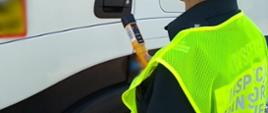 Inspektor ITD w odblaskowej kamizelce stoi obok kabiny zatrzymanej do kontroli ciężarówki i trzyma w ręku alkomat przesiewowy.