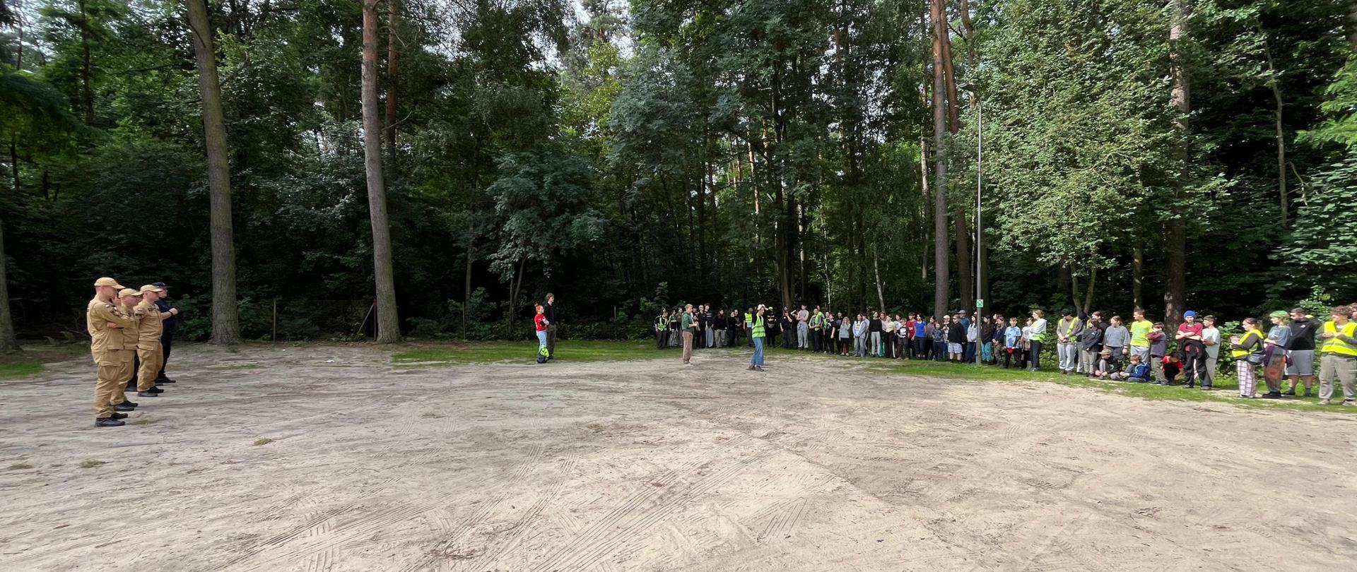 Apel na obozie harcerskim w lesie. Strażacy i policjanci stoją po lewej stronie dużej, piaszczystej polany, a naprzeciwko nich ustawiony jest szereg młodzieży i opiekunów.