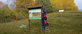 Tablica informująca o obszarze Natura 2000 Murawy Gorzowskie oraz dwóch mężczyzn montujących daszek nad tablicą.