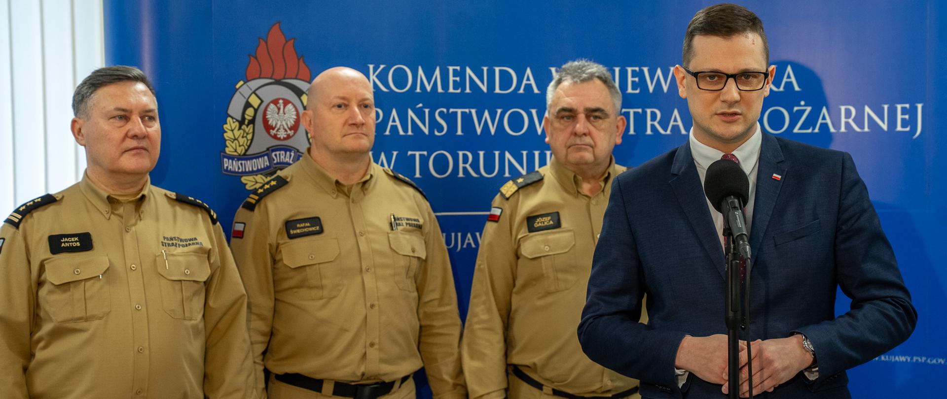 Wojewoda Kujawsko-Pomorski Michał Sztybel przemawia podczas briefingu prasowego w towarzystwie komendantów Państwowej Straży Pożarnej ubranych w mundury służbowe. W tle widoczne są symbole i logotypy straży pożarnej.