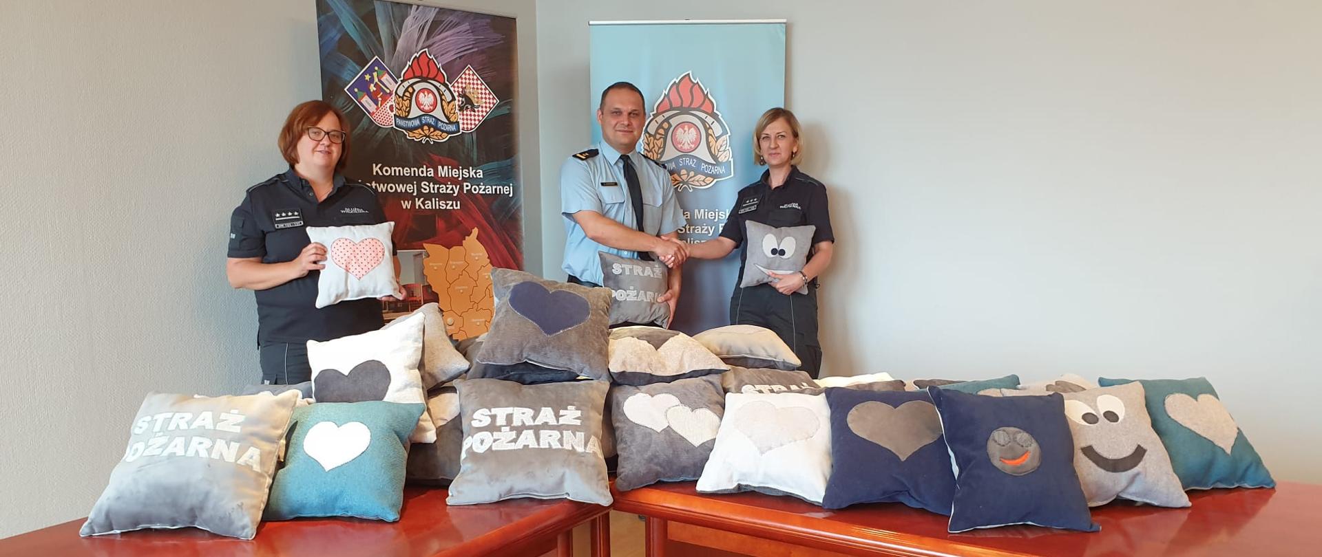 Na zdjęciu dwie funkcjonariuszki Służby Więziennej. W środku, w niebieskiej koszuli Komendant Miejski PSP w Kaliszu bryg. Sławomir Kotoński. Komendant ściska dłoń jednej z pań. Za plecami funkcjonariuszy banery promocyjne kaliskiej komendy z logo PSP i powiatu. Przed osobami stół drewniany, brązowy, na którym ułożone są poduszki. Pomieszczenie ma jasno żółte ściany.