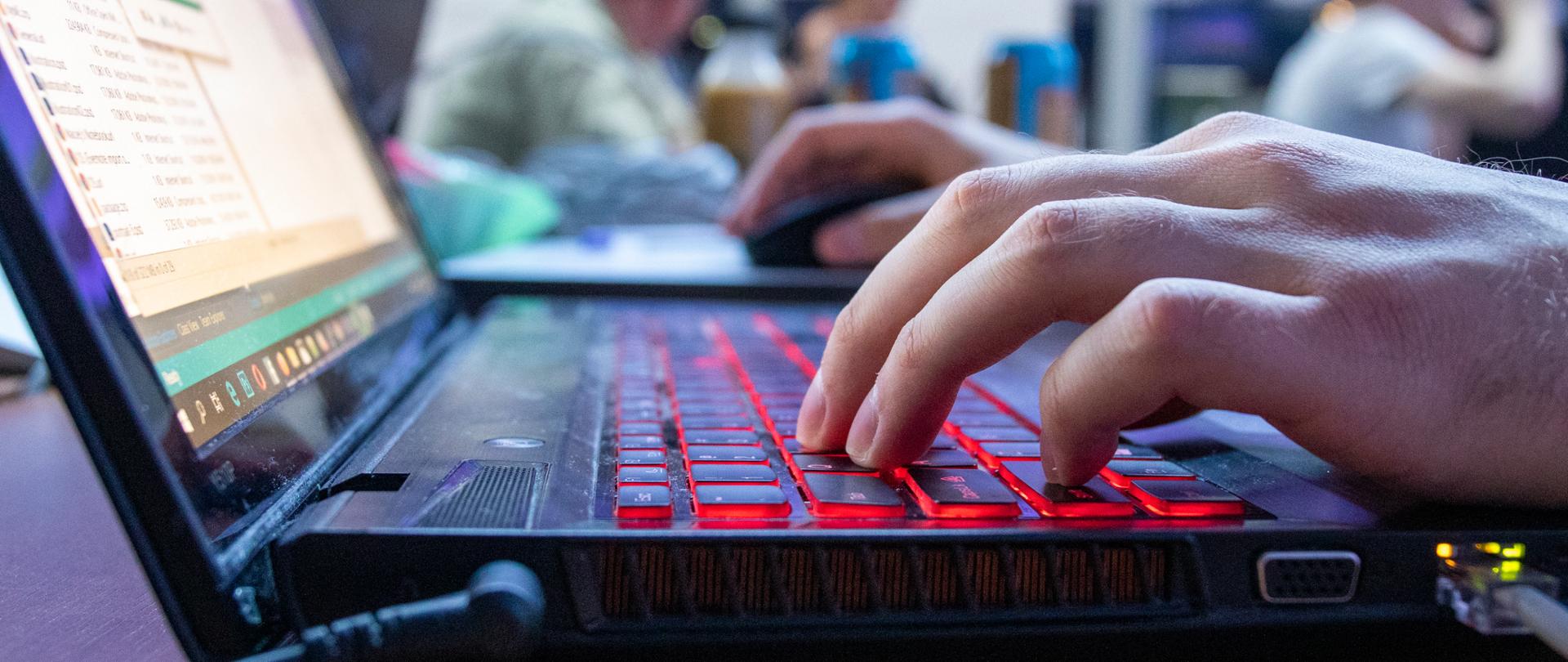 zdjęcie przedstawia klawiaturę laptopa i ręce programisty na tle innych uczestników hackhatonu