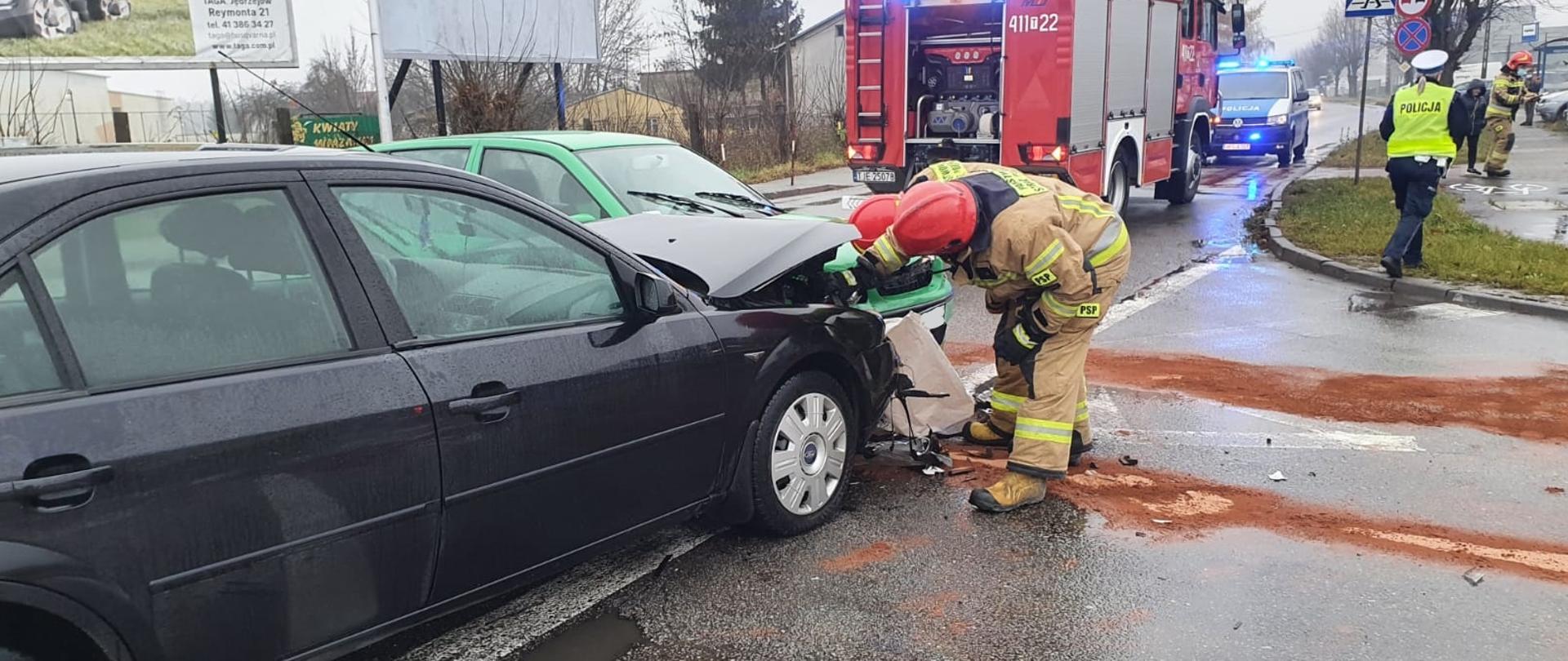 Zdjęcie przedstawia dwa rozbite pojazdy osobowe na środku skrzyżowania. Przy jednym z nich pracują strażacy w umundurowaniu specjalnym, którzy odłączają akumulator. W tle samochód ratowniczo-gaśniczy oraz radiowóz policyjny. 