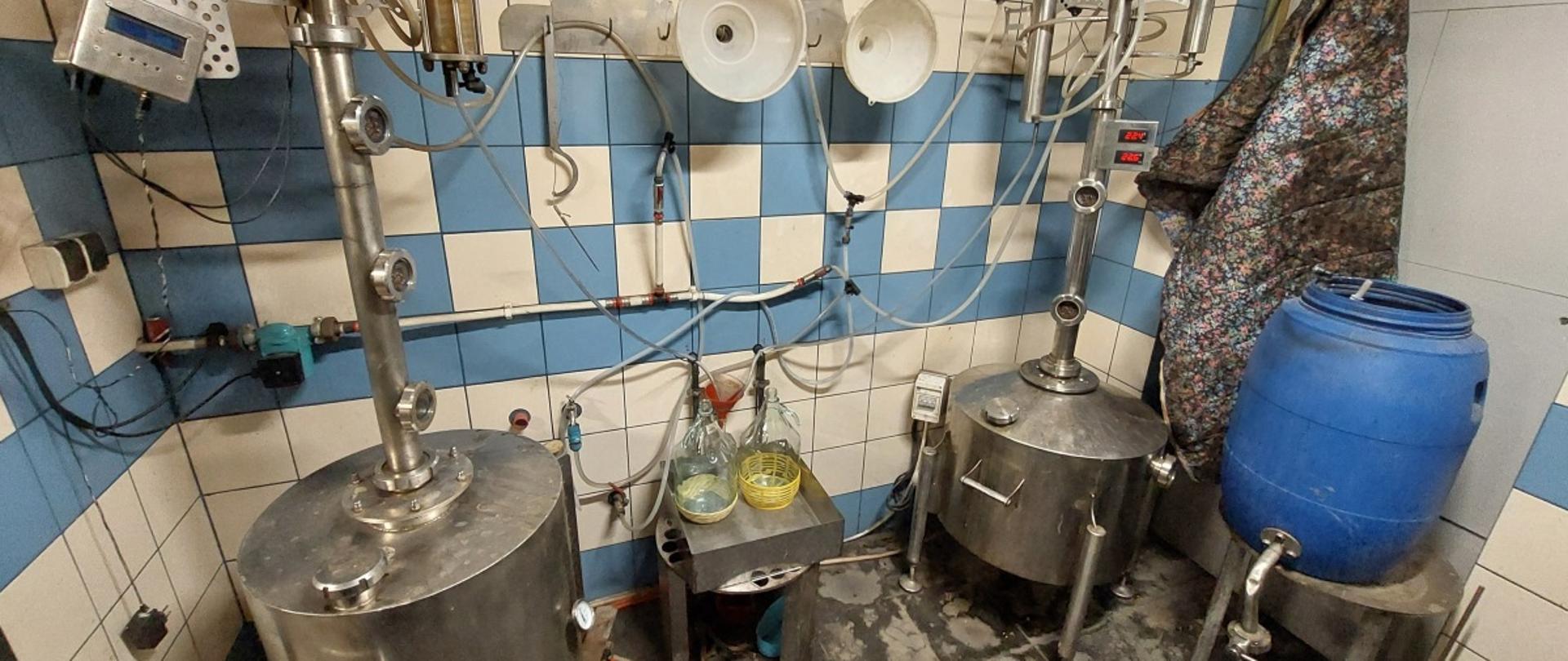 Pomieszczenie techniczne z metalowymi zbiornikami połączonymi rurami i przewodami, obok stoi niebieski plastikowy pojemnik oraz mały stół z naczyniami.