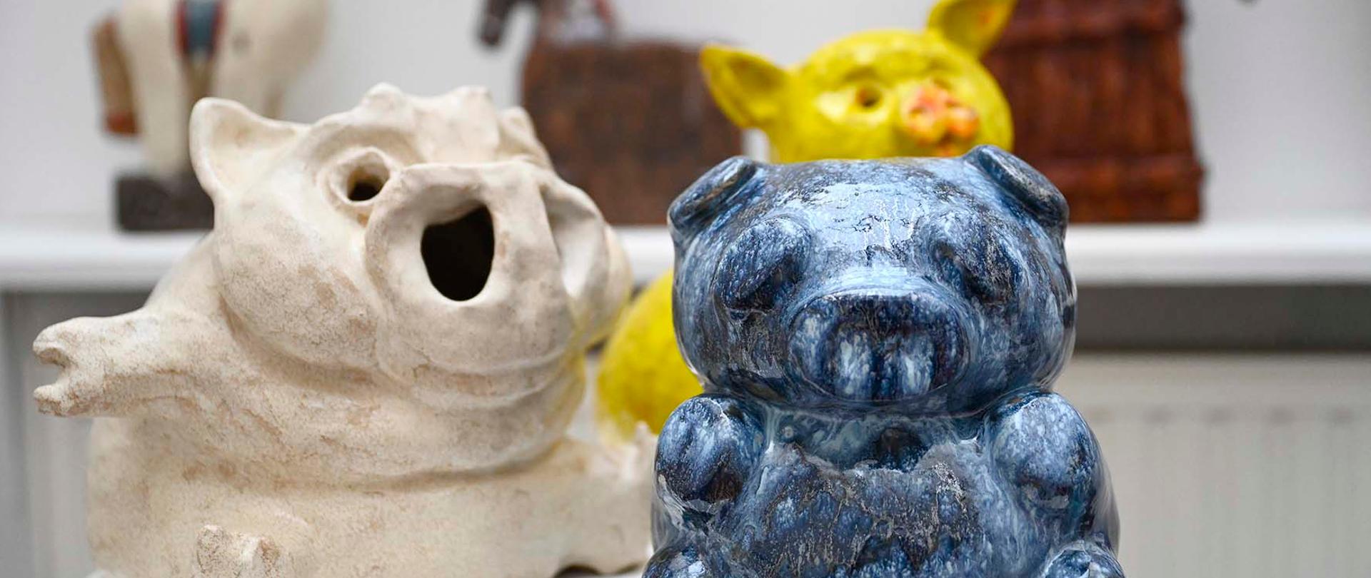 Na zdjęciu widać kilka ceramicznych figurek zwierząt, m.in. niebieską świnkę na pierwszym planie oraz jasnego hipopotama i kolorowe figurki koni w tle.