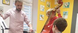 Na zdjęciu znajdują się pedagog prowadzący warsztaty - Wojciech Front oraz uczeń z kontrabasem i uczeń słuchający, w sali rytmicznej PSM w kolorach szaro, żółty, biały, na ścianach wiszą kolorowe obrazki muzyczne