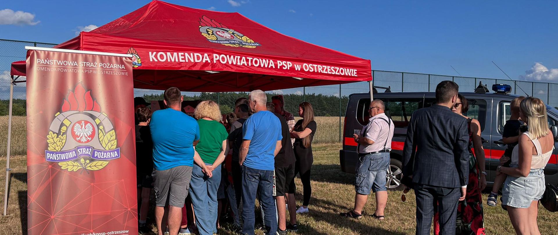 Grupa zainteresowanych osób prelekcją na temat bezpieczeństwa stoi tyłem do fotografa. Słuchacze stoją przy czerwonym namiocie z logiem Państwowej Straży Pożarnej i napisem "Komenda Powiatowa PSP w Ostrzeszowie". Po lewej stronie baner z logiem Państwowej Straży Pożarnej. Na drugim planie samochód typu bus straży pożarnej.