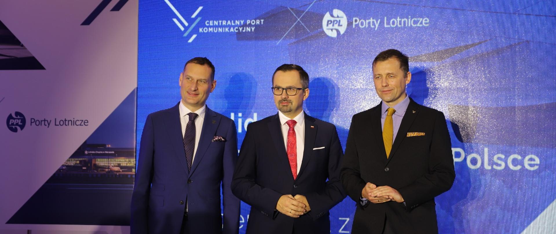 Trzy osoby stoją w jednym rzędzie obok siebie. W środku wiceminister Marcin Horała.