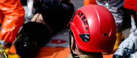 Strażak w ubraniu specjalnym w hełmie patrzy jak strażacy w ubraniach chemicznych aparatach ochrony dróg oddechowych udzielają pierwszej pomocy w namiocie dekontaminacyjnym.