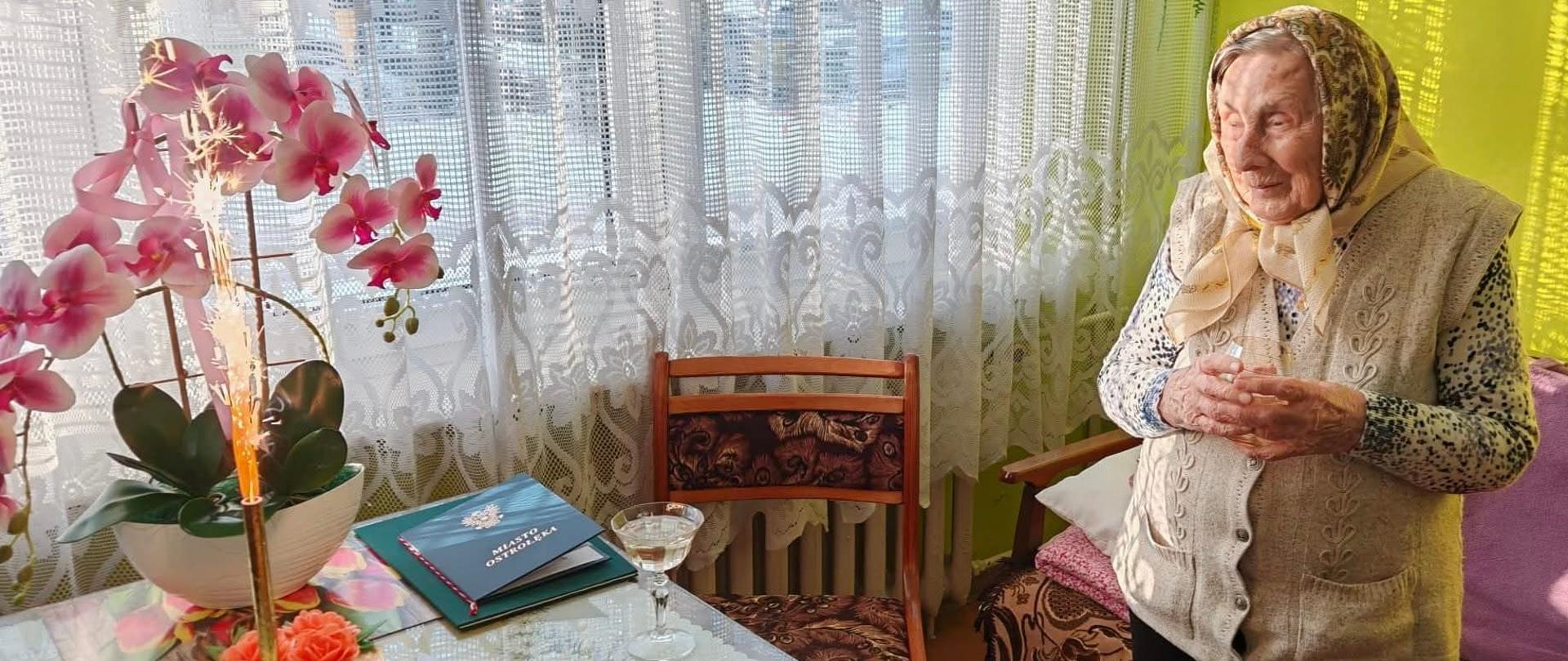 Starsza kobieta stoi przy stole w jasnym pokoju. Na stole stoi tort z zapaloną fontanną iskier oraz kwiat doniczkowy – różowy storczyk. Obok leży zielony dyplom z logo KRUS. Kobieta ma złożone dłonie i patrzy w stronę tortu. Wnętrze jest ciepłe i domowe.