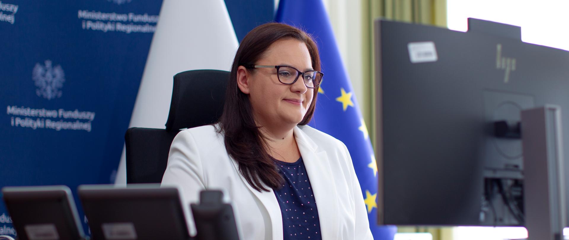 Na zdjęciu wiceminister funduszy i polityki regionalnej Malgorzata Jarosińska-Jedynak siedząca przed komputer w trakcie wideokonferencji podczas Smart City Innovation Ecosystems Festival. W tle flaga Polski i Unii Europejskiej. 