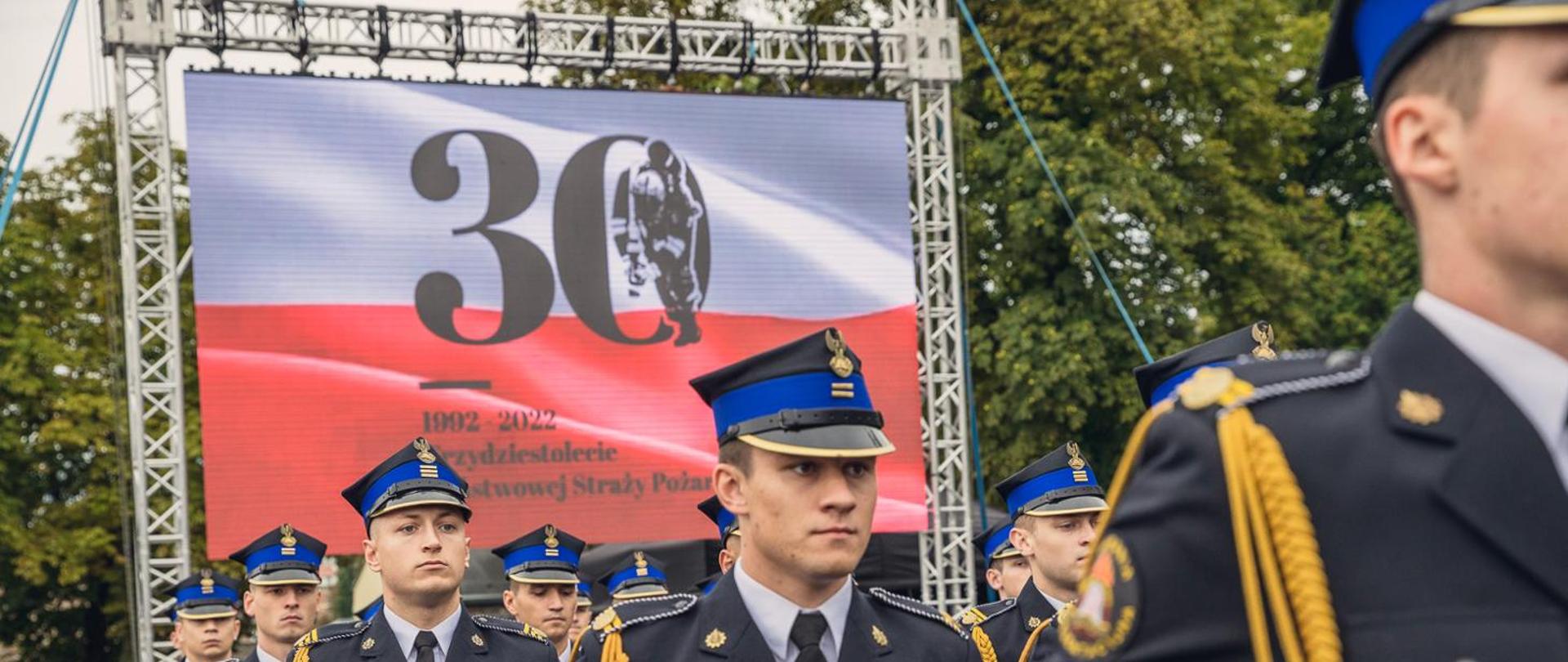 Maszeruje kompania reprezentacyjna, za nimi na telebimie wyświetlona flaga RP z napisem okolicznościowym 30 lecia PSP