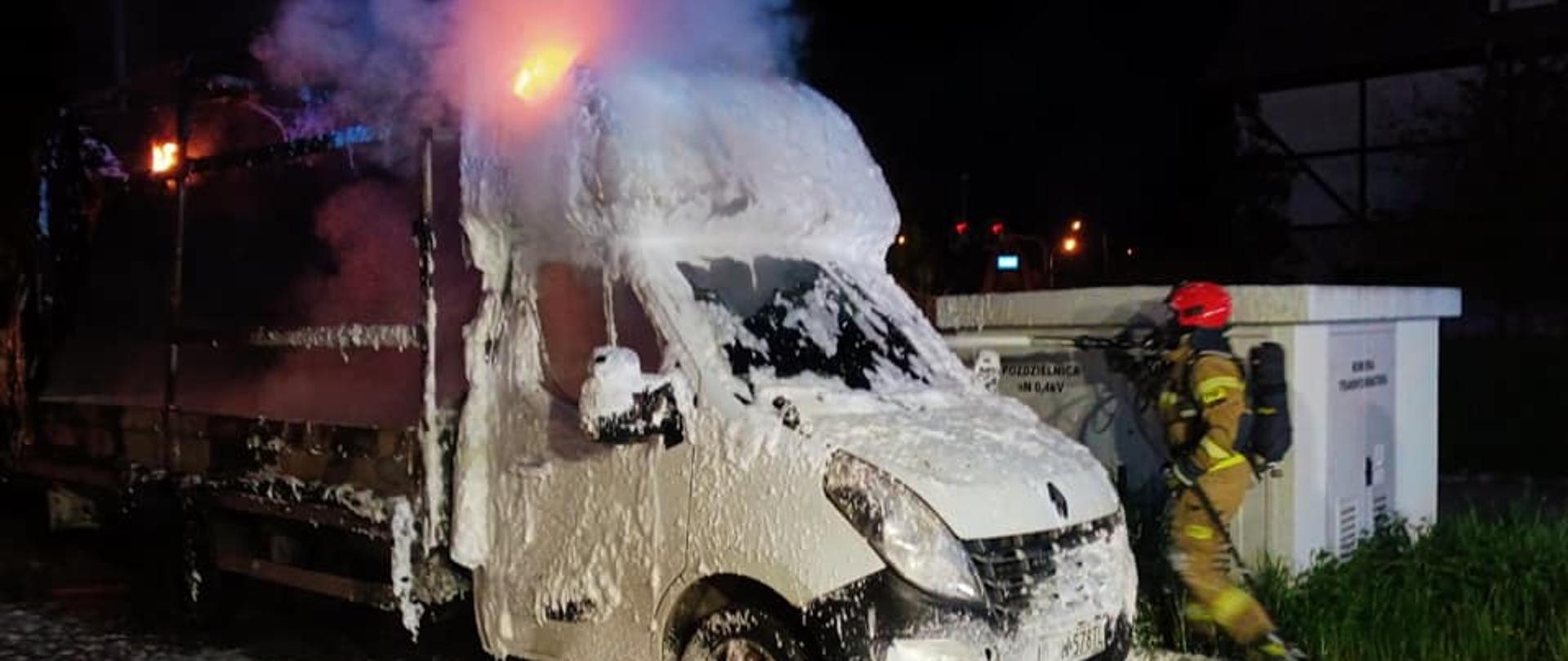 Zdjęcie przedstawia strażaka, który gasi pianą palący się samochód dostawczy.