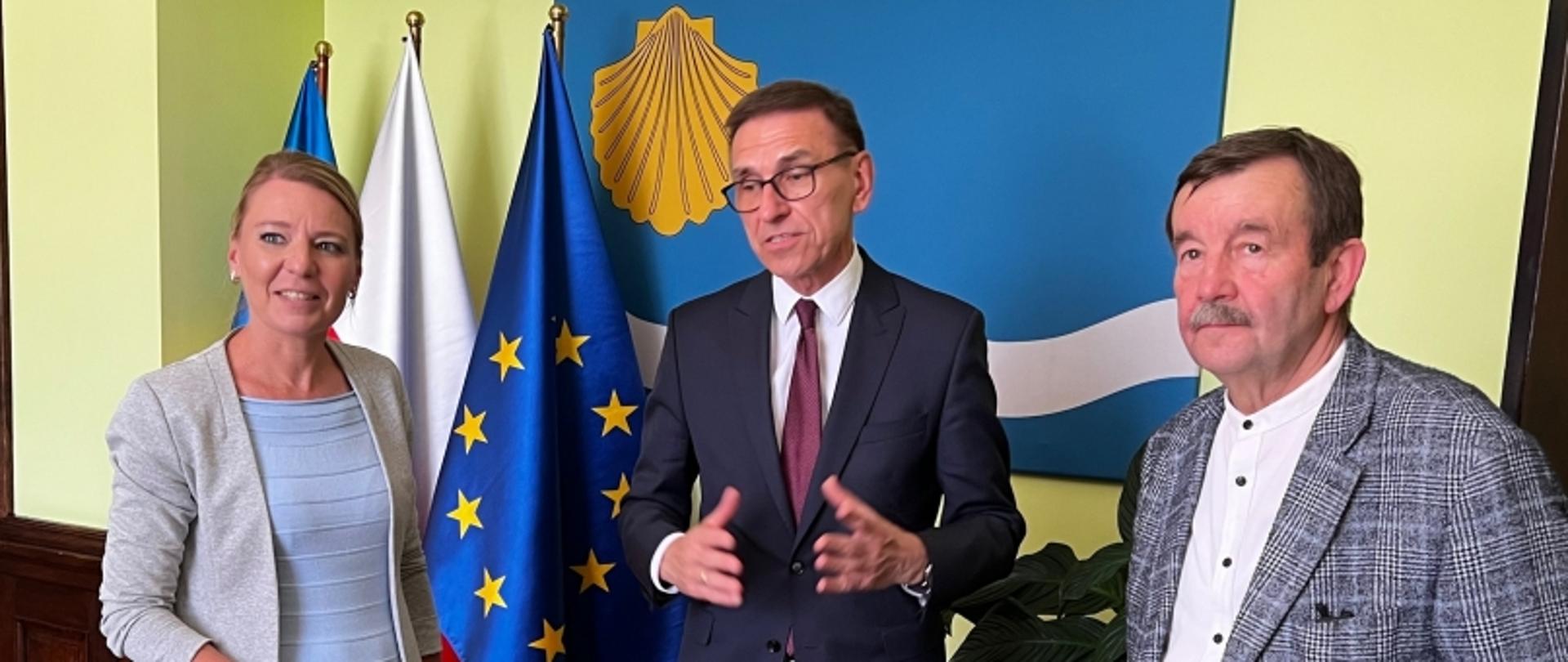 Trzy osoby na tle flag Polski, UE i flagi z żółtą muszlą, z drewnianym panelem i roślinami w tle.