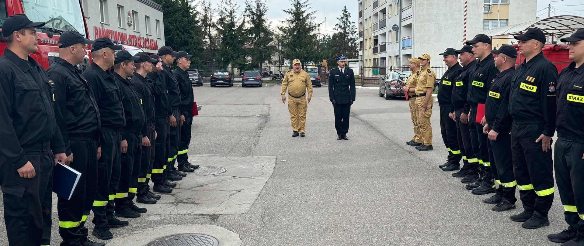 Dwie zmiany służbowe stoją naprzeciwko siebie. Po środku stoją Komendant Powiatowy PSP oraz Zastępca Dowódcy JRG