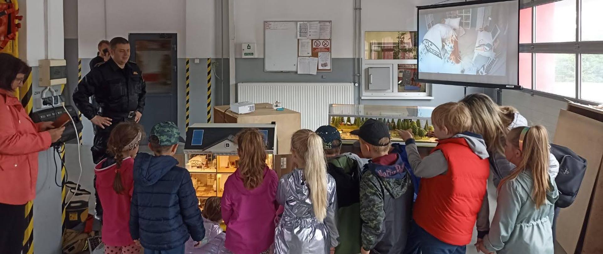 Pokazy dla dzieci przy użyciu Mobilnego Symulatora Zagrożeń Pożarowych w KP PSP Chełmno. Zdjęcie wykonano w garażu bojowym komendy, znajdują się na nim dzieci podczas pokazów. 