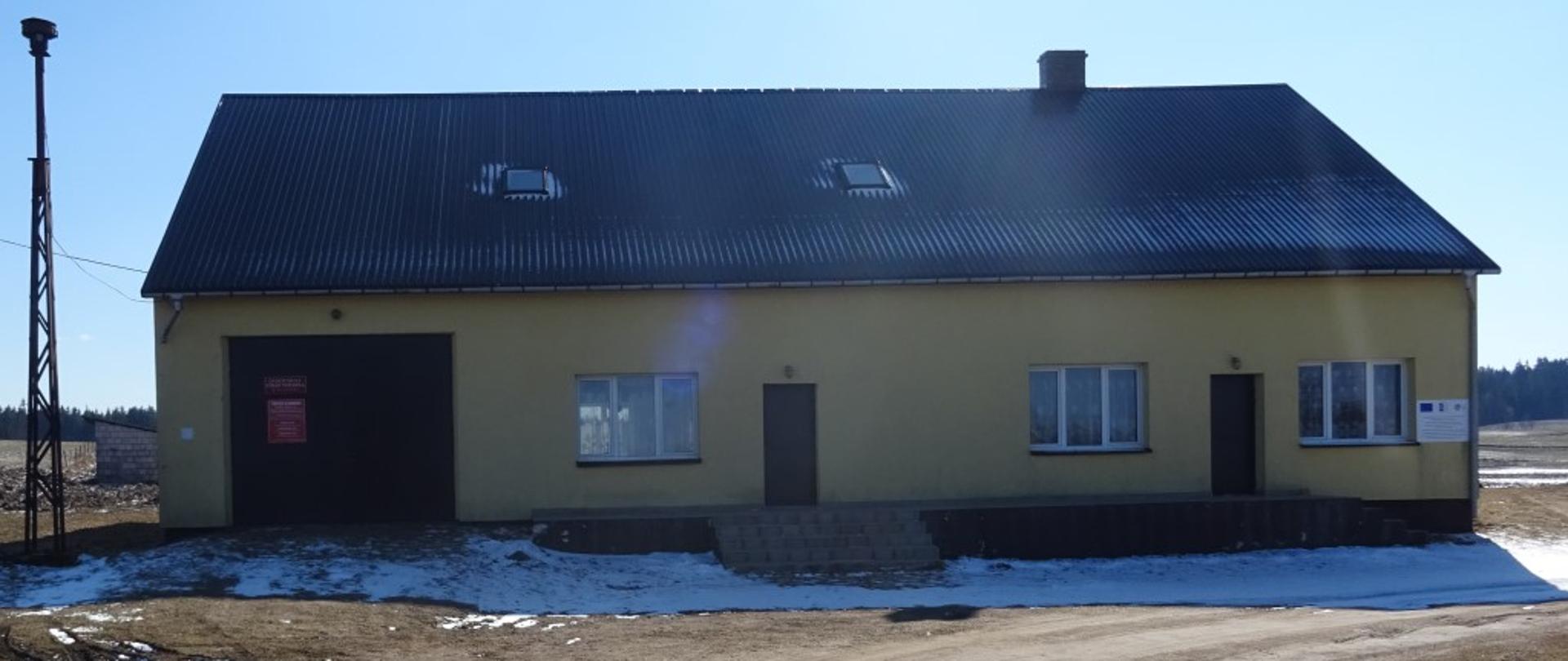 Budynek koloru żółtego ,brama garażowa koloru brązowego. Dwoje drzwi do budynku od frontu. Dach kryty blacha koloru brązowego. Podjazd wysypany ziemią. 