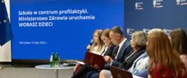 Przedstawiciele Ministerstwa Edukacji Narodowej i Ministerstwa Zdrowia siedzą w rzędzie. W tle za nimi wyświetlana jest prezentacja z tytułem: Szkoła w centrum profilaktyki. Ministerstwo Zdrowia uruchamia WOBASZ DZIECI.