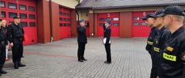 Zastępca Komendanta Powiatowego Państwowej Straży Pożarnej w Rypinie Pan brygadier Andrzej Górecki oddaje honor Komendantowi Powiatowemu Państwowej Straż Pożarnej w Rypinie, Panu starszemu brygadierowi Grzegorzowi Łydkowskiemu. Za nimi czerwone wrota garażowe. Po prawej i lewej stronie na polbruku stoją strażacy w dwuszeregach. 