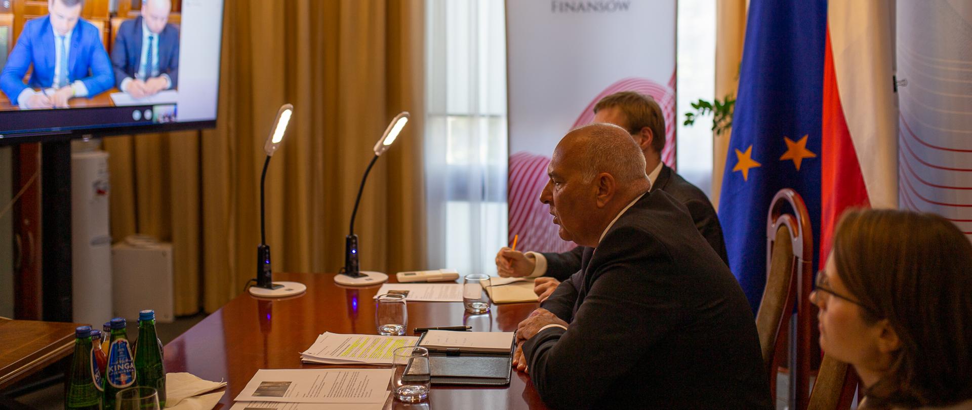 Minister Tadeusz Kościński, wiceminister Jan Sarnowski i szefowa KAS Magdalena Rzeczkowska podczas telekonferencji ze stroną ukraińską, siedzą przy stole, przed nimi telebim, w tle flagi Polski i UE