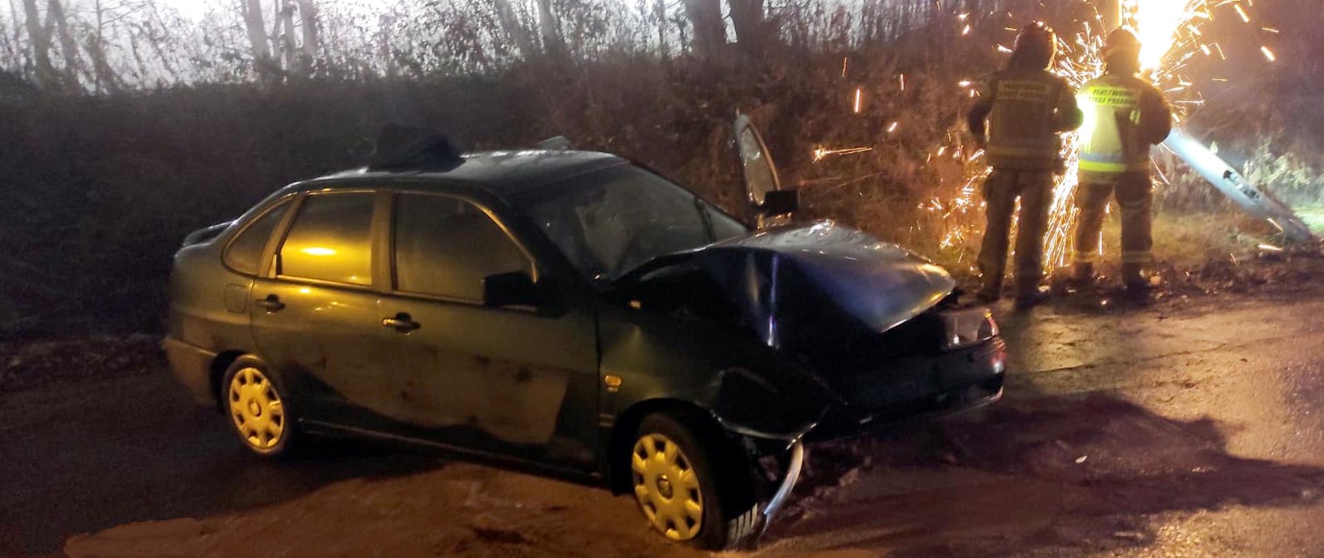 Zdjęcie przedstawia stojący na poboczu samochód marki Seat z rozbitym przodem. Obok dwaj strażacy usuwają przy pomocy pilarki uszkodzoną przez pojazd latarnię. Z miejsca cięcia sypią się iskry. Z tyłu widać zarośla.