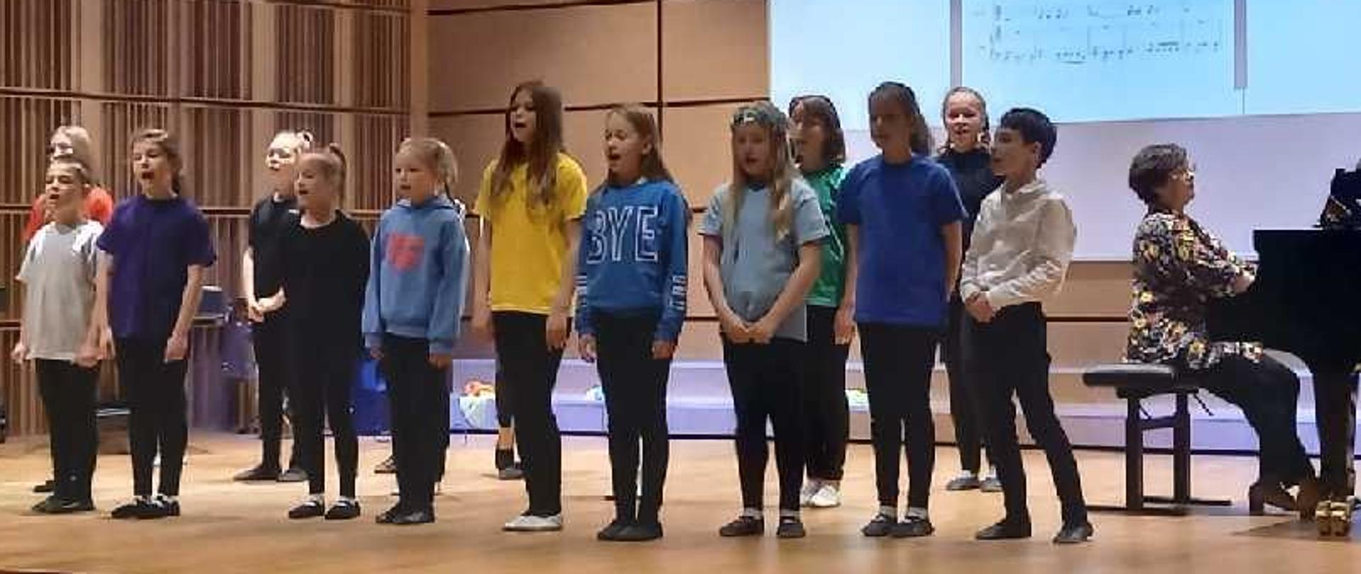 Zdjęcie przedstawia uczniów śpiewających, stojących w dwóch rzędach. Obok nich nauczycielka grająca na fortepianie.