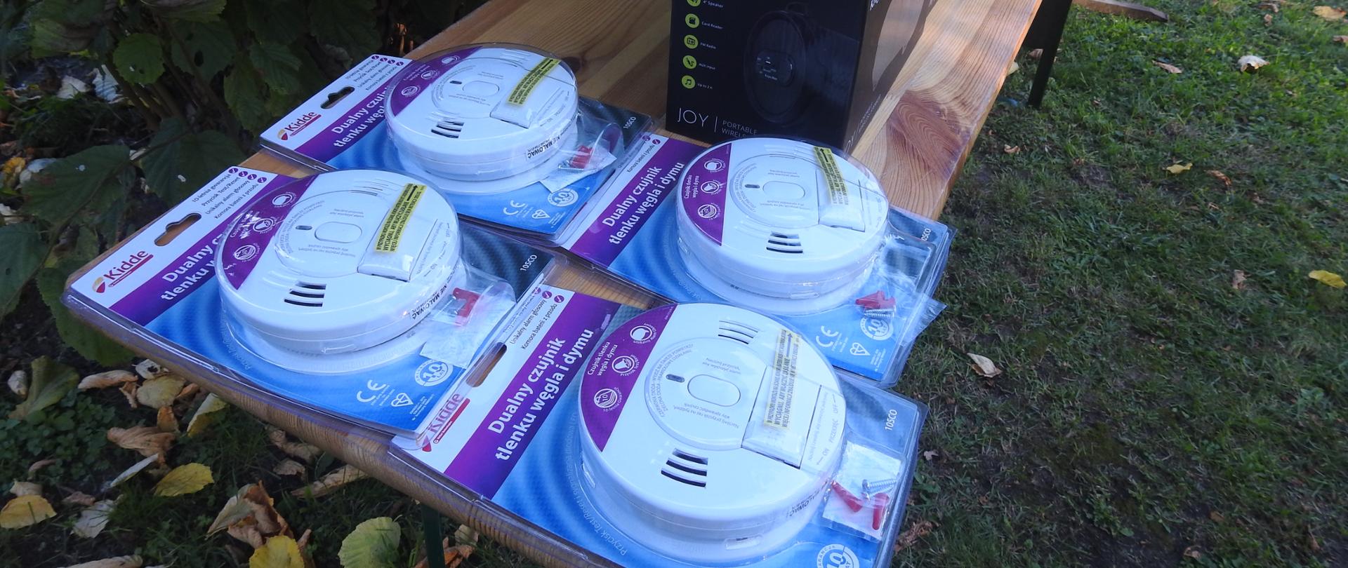 Cztery czujniki czadu leżą na stoliku. Stolik stoi na trawie. Na trawie leż liście. 