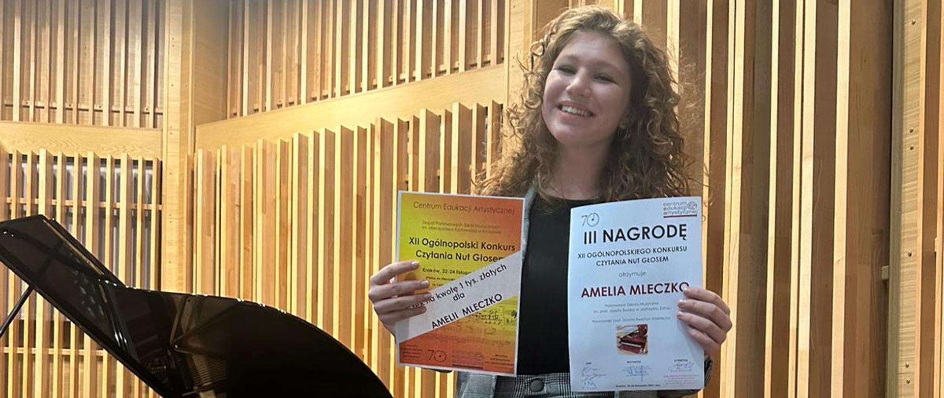 Elegancko ubrana nastolatka stoi przed fortepianem i trzyma w rękach dwa dyplomy.