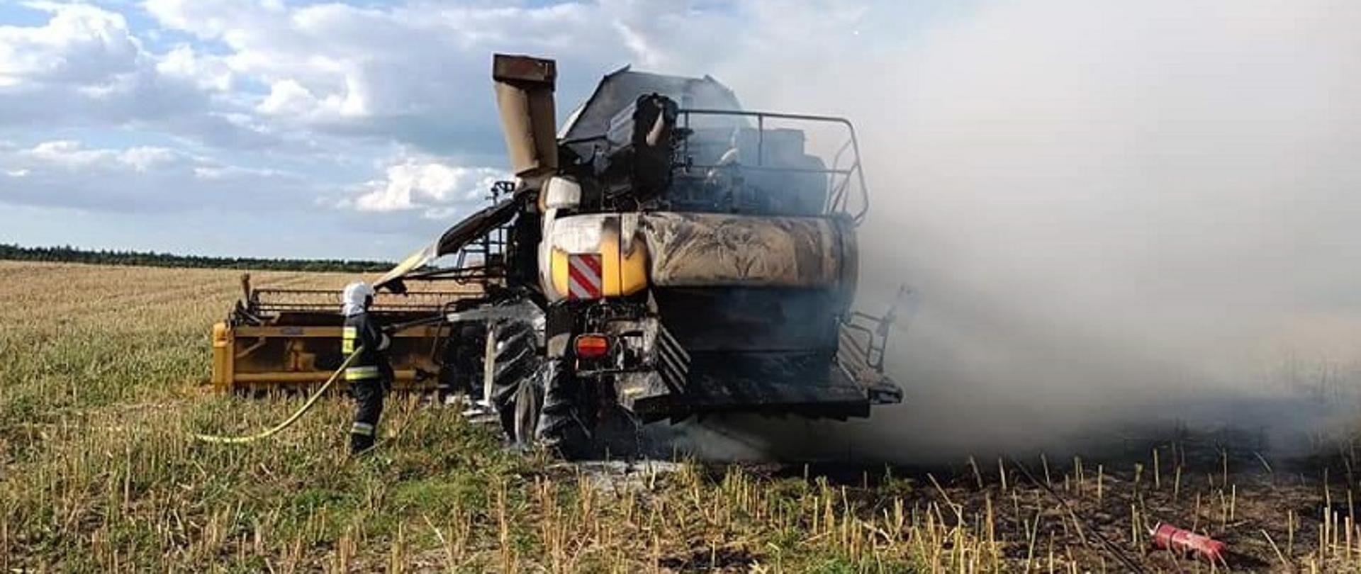 Na środku pola stoi spalony kombajn, po lewej stronie stoi strażak. Nad maszyna unoszą się kłęby białego dymu.
