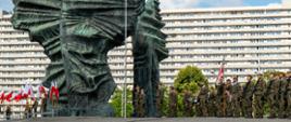 Pomnik Powstań Śląskich w Katowicach, pod pomnikiem stojąca kompania wojskowa wraz z pocztem sztandarowym, z drugiej strony biało czerwone flagi RP, w tle blok mieszkalny 
