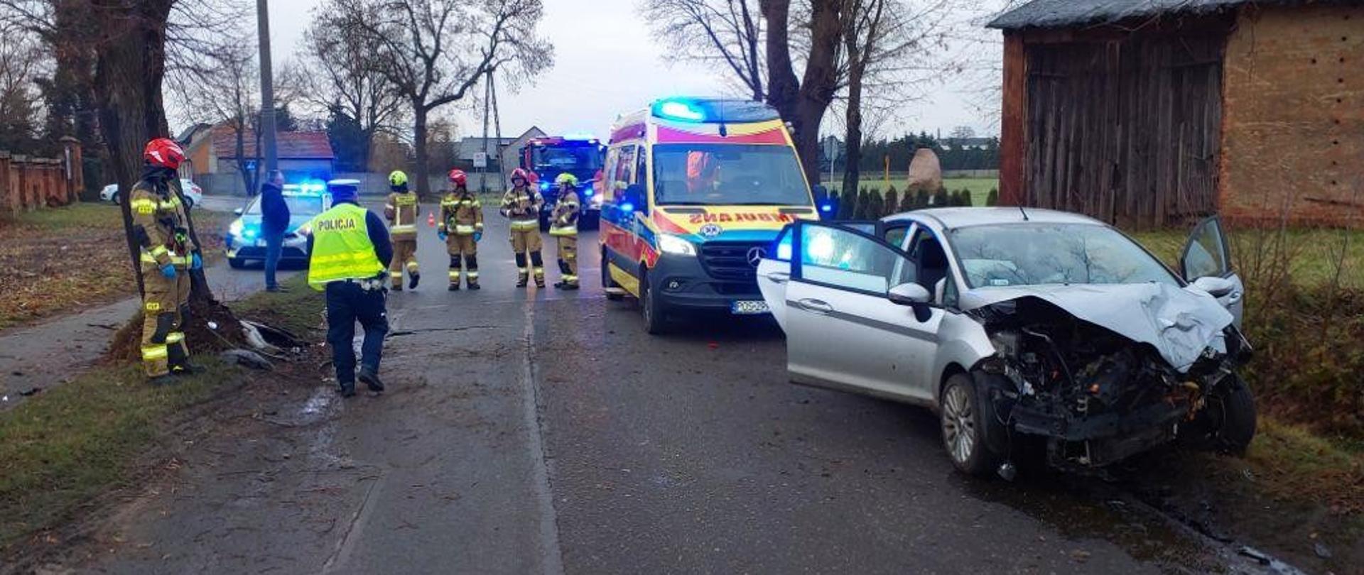 Wrak rozbitego pojazdu, w tle strażacy oraz ambulans