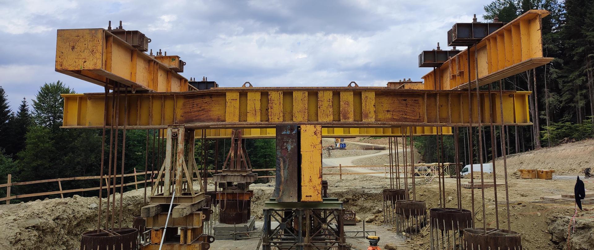 Widok z poziomu ziemi na budowę estakady i specjalną konstrukcję stalową do próbnego obciążenia. Sprawdzenie nośności wykonano metodą belki odwróconej. Na pierwszym planie widać mierniki stojące na statywach. 