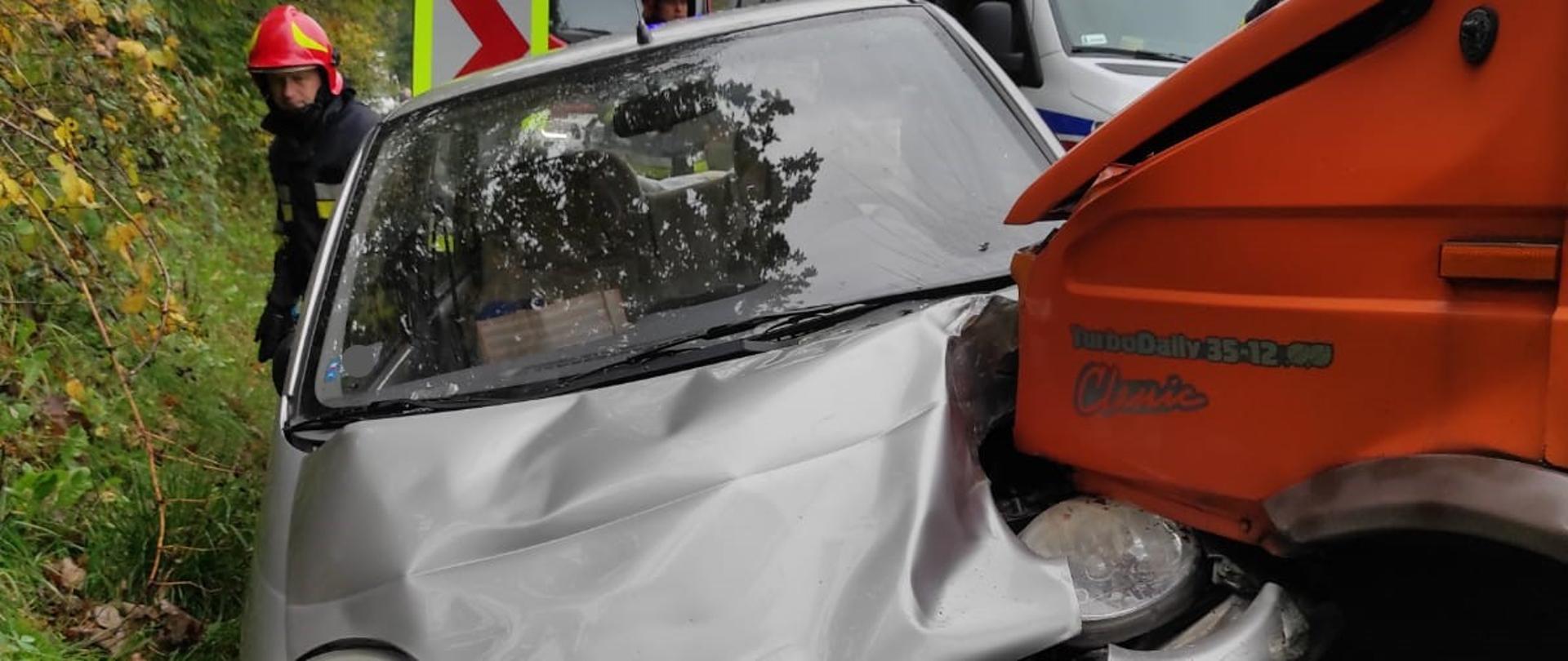 Zdjęcie przedstawia samochód dostawczy i osobowy, które zderzyły się i w następstwie tego znalazły się częściowo poza jezdnią. Na drugim planie częściowo widoczna jest karetka pogotowia i samochód straży pożarnej oraz strażacy. W tle widoczne drzewa i zarośla.