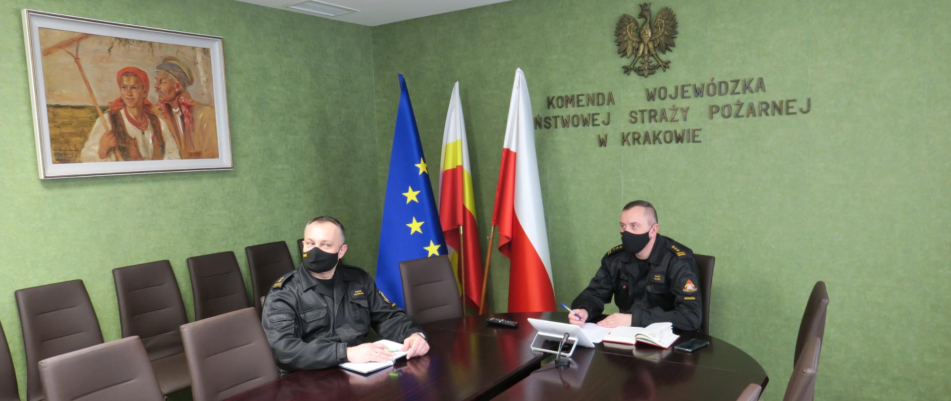 Sala konferencyjna KW PSP Kraków. W centralnej części duży stół przy którym siedzi Komendant i Zastępca. Ubrani w czarne ubrania dowódcze i maski zakrywające usta i nos.