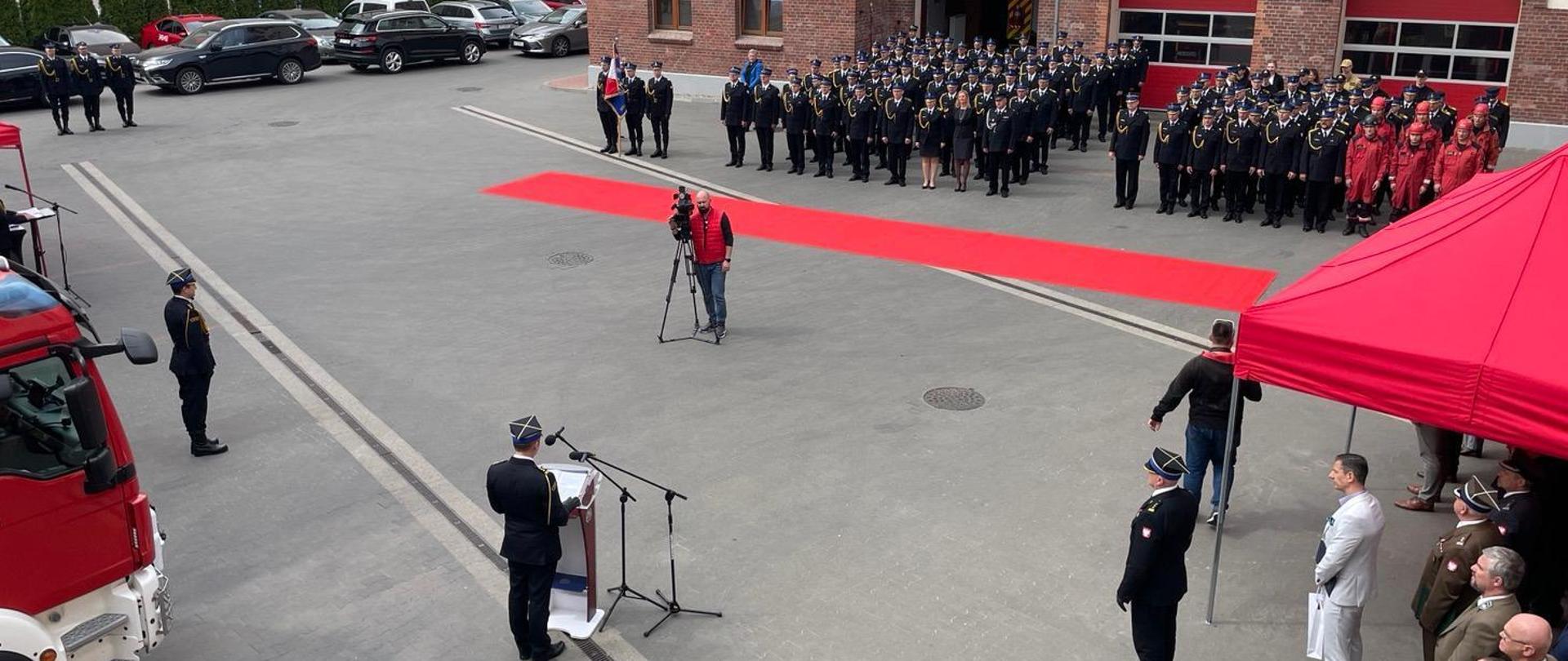 Widok z góry na plac uroczystości, w środku operator z kamerą. Z lewej przód pojazdu pożarniczego, w tle pododdziały awansowanych. 