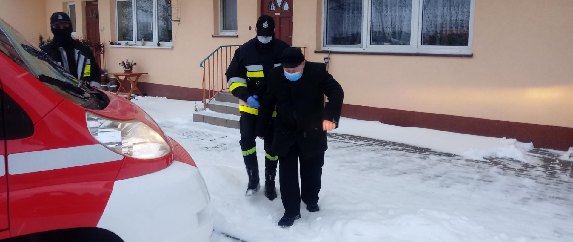 Strażak pomaga iść mężczyźnie do samochodu strażackiego. w tle jednopiętrowy beżowy budynek. na ziemii śnieg.