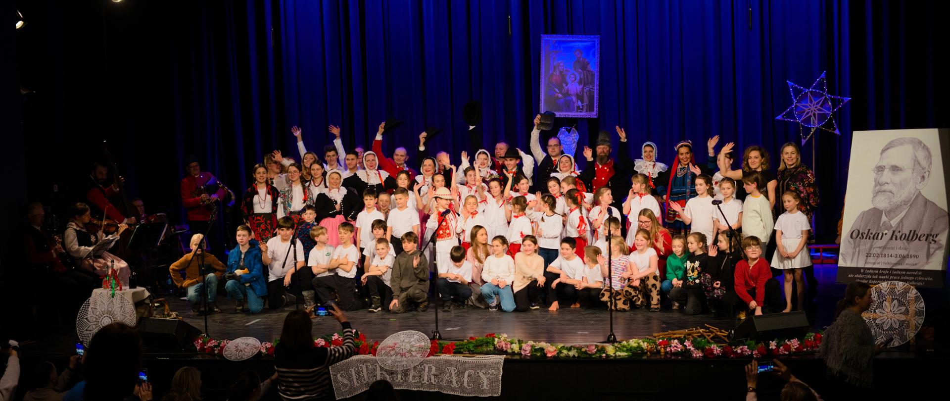 Grupa dzieci i dorosłych na scenie. Dorośli w tradycyjnych strojach ludowych. W tle nad ich głowami obraz z motywem religijnym. Scena przyozdobiona kwiatami oraz dekoracyjnymi koronkami. Po lewej stronie zespół smyczkowy. Po prawej stronie czarno - biały portret Oskara Kolberga. 