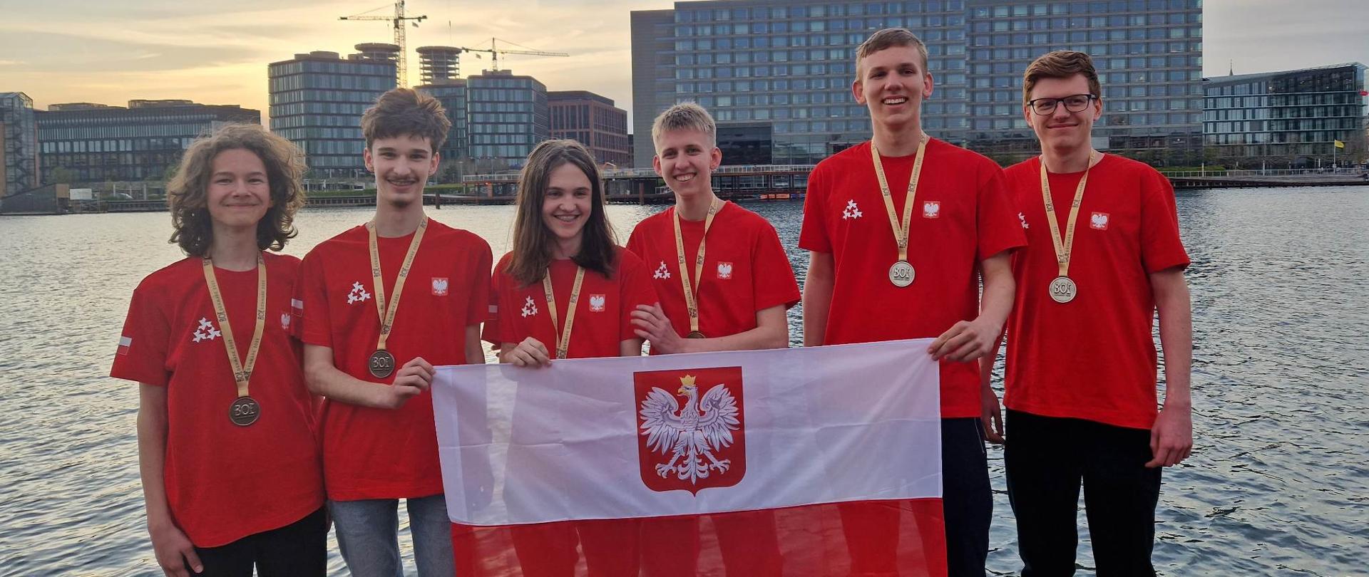 Grupa młodych ludzi ubranych w czerwone koszulki i trzymających polską flagę. W tle morze i wysokie szklane budynki 