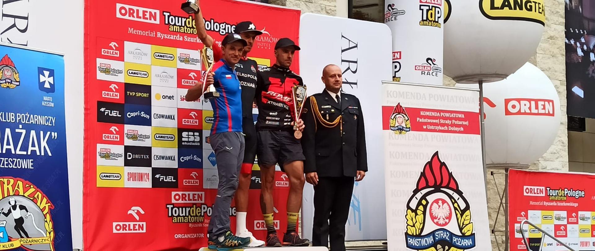 Zdjęcie przedstawia trzech kolarzy, którzy stoją na podium po zakończonym biegu. Obok stoi strażak w ubraniu galowym. Strażacy ukończyli wyścig Tour de Pologne Amatorów 2022