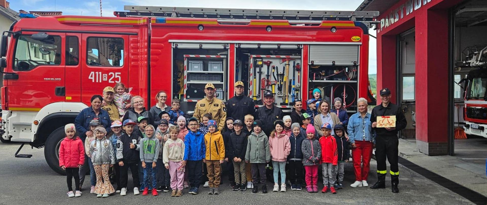 Kolorowa fotografia wykonana na zewnątrz w pogodny dzień. Przedstawia grupę przedszkolaków, które odwiedziły strażnicę PSP oraz strażaków w ubraniach służbowych na tle samochodu pożarniczego.